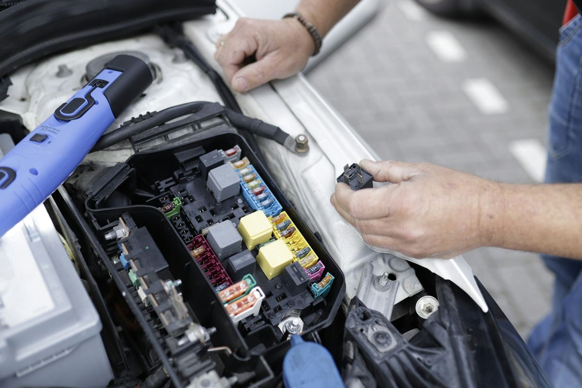 Twee handen werken aan de zekeringkast onder de motorkap, met een meettoestel ernaast